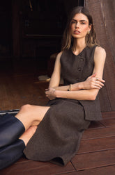 A woman is sitting on a wooden floor, leaning against a wooden wall. She is wearing a sleeveless, dark-colored vest with buttons and a matching knee-length skirt. She has gold jewelry, including a bracelet, ring, and earrings, and is wearing black knee-high boots.
